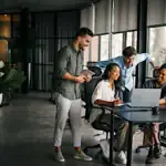 Ein Team aus vier Personen arbeitet gemeinsam im Büro an einem Laptop und bespricht ein Projekt zur Unternehmensgründung.